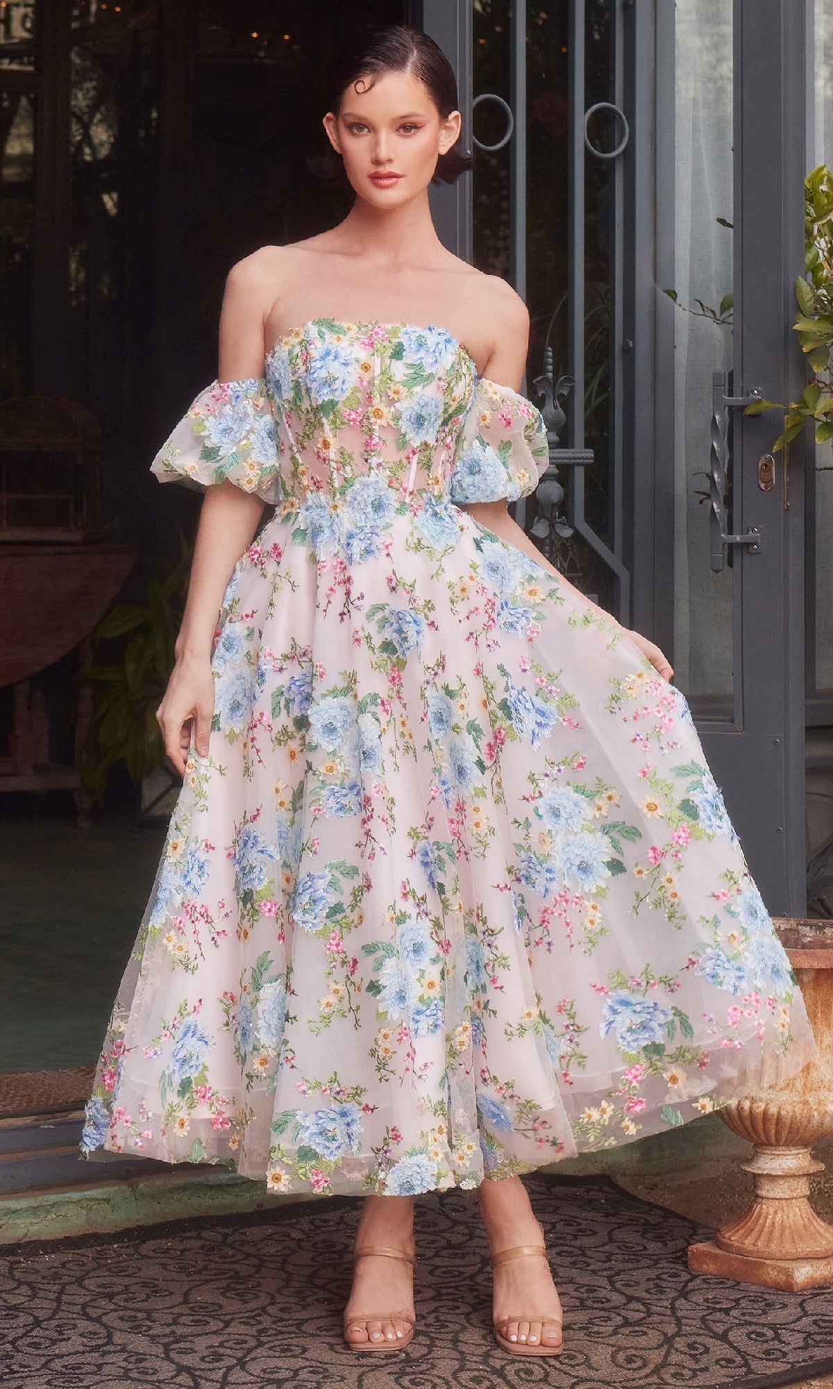 A woman stands outside by a glass door wearing the Andrea & Leo A1406 Floral Prom Dress, featuring a strapless corset bodice and removable puff sleeves, paired with nude heels.